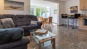 A modern living room with a dark grey sofa, a glass coffee table, and a dining area with wicker chairs. A kitchen with a stove and bar stools is visible in the background. - Wohnung in St.peter Ording (St. Peter-Ording)