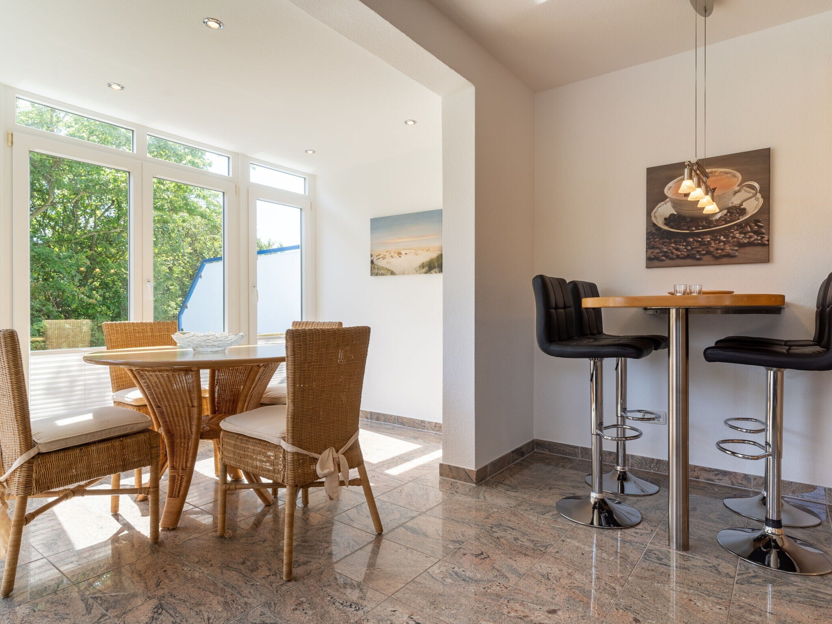A dining area with wicker chairs and a table, a bar area with stools, and large windows with a view of greenery.