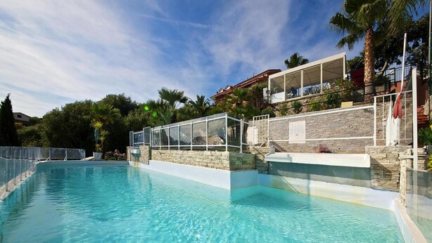 Una piscina limpida con parete in pietra e ringhiera in vetro, immersa nel verde e affiancata da un edificio con balcone.
