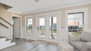 A spacious living room with a grey sofa, a wooden staircase, and a view of the outside. - A New Hope: Ocean View with Private Pool and Easy Beach Access (Oak Island)