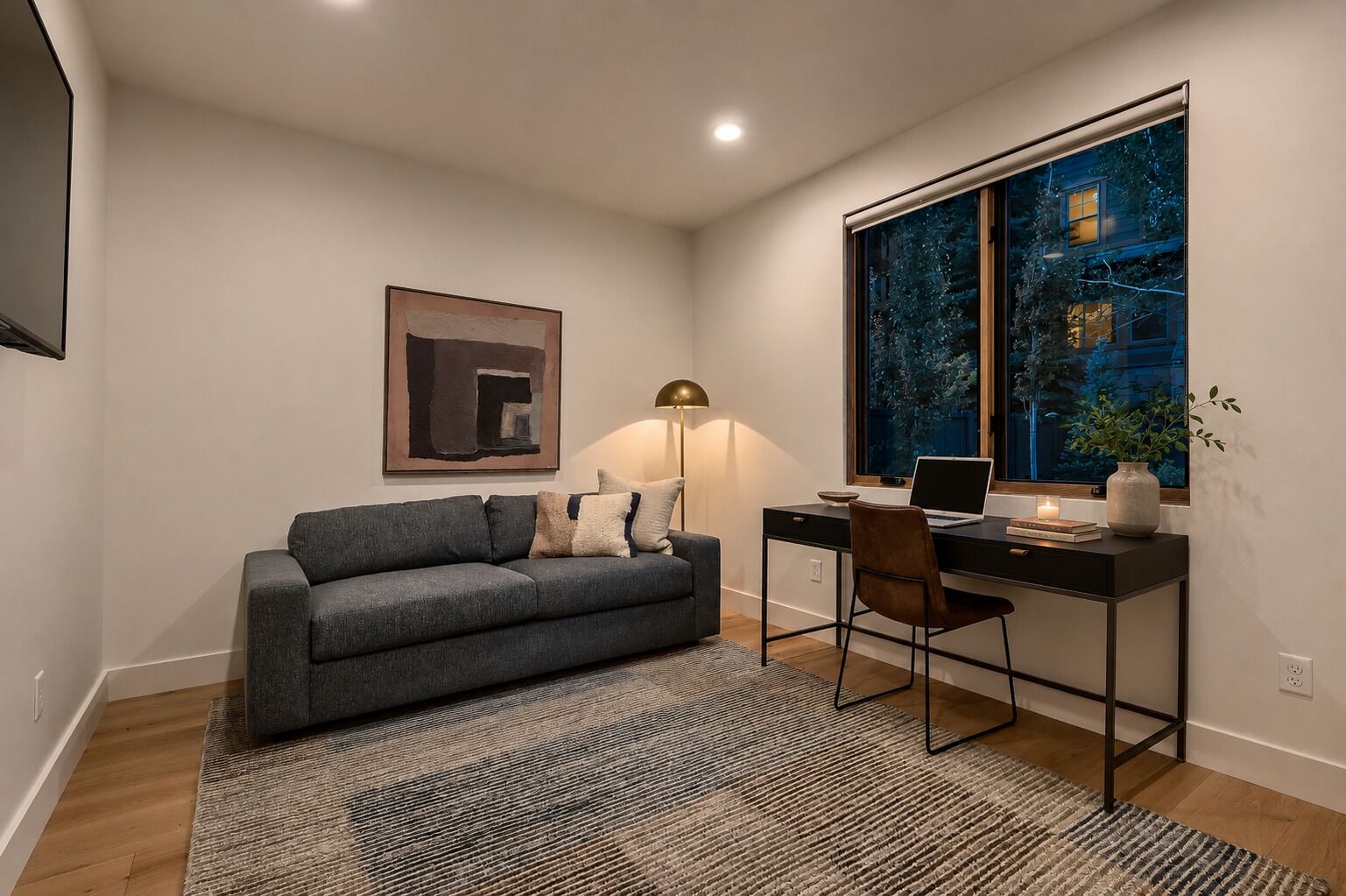 Une chambre moderne avec un canapé gris, un bureau avec un ordinateur portable, une grande fenêtre donnant sur le paysage nocturne et un tableau abstrait encadré accroché au mur.