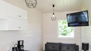 A modern kitchen with white cabinets, a black kettle, and a coffee maker. There is a grey sofa and a round table with a remote control. A TV is mounted on the wall above the sofa. - Bungalow in Kolczewo Near Baltic Sea Beach (gmina Wolin)