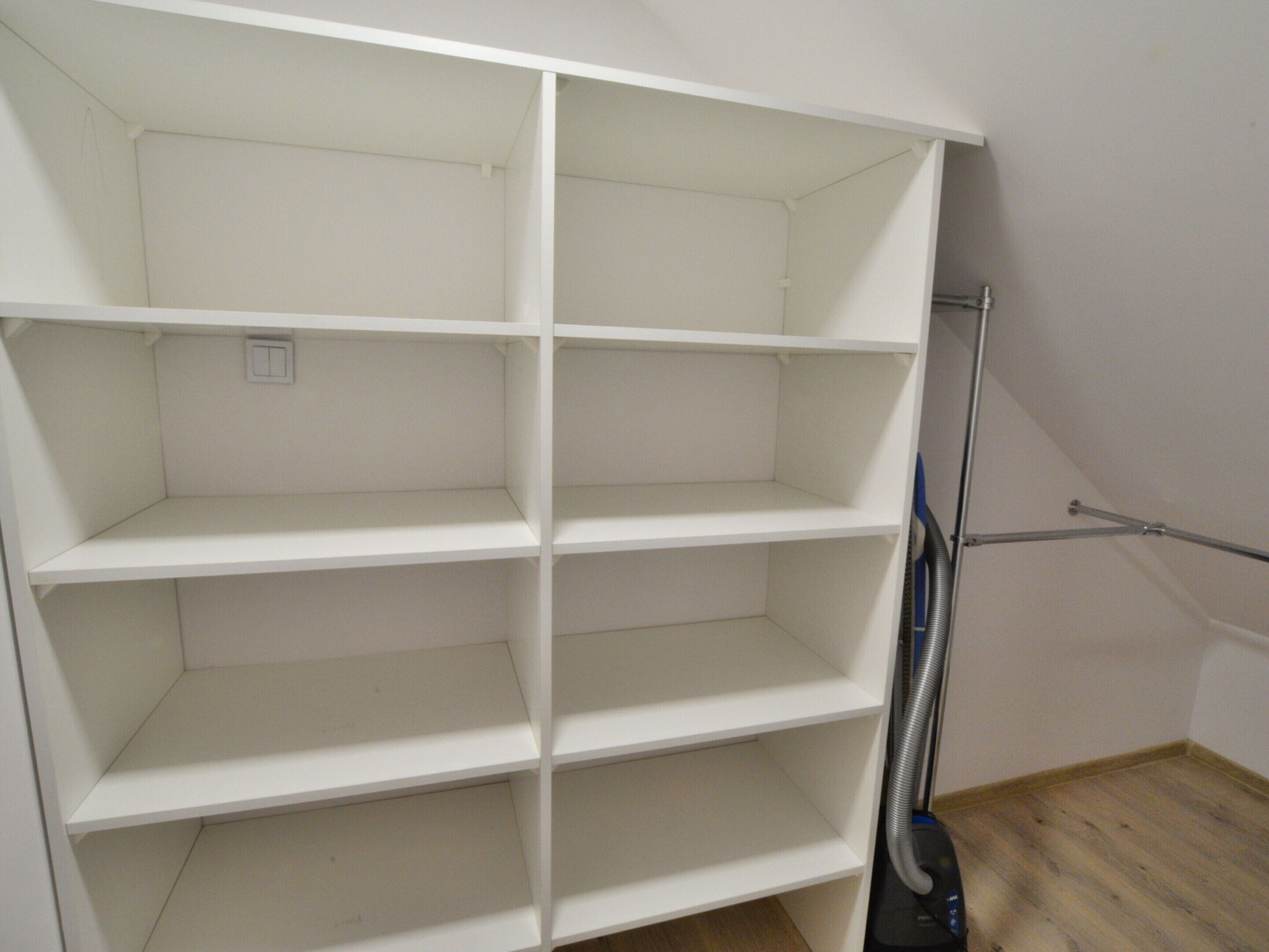 A white, empty storage unit with multiple shelves, a vacuum cleaner, and a wooden floor.