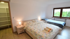 A bedroom with a wooden floor, a bed with floral bedding, two bedside tables with lamps, and a window with a view of greenery. - Spacious Holiday Apartment With a Large Terrace (Kołobrzeg)