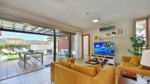 Una sala de estar moderna con un televisor grande, sofás amarillos, una zona de comedor y un ventanal corredero a un patio.