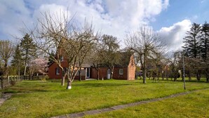 A brick building with a red door and a small garden area with grass and trees. - Ferienhaus in Garding (Garding)