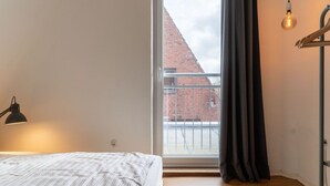 A bedroom with a bed, a desk lamp, a hanging light bulb, and a view of a brick building through a window with a curtain. - Ferienhaus in Garding (Garding)