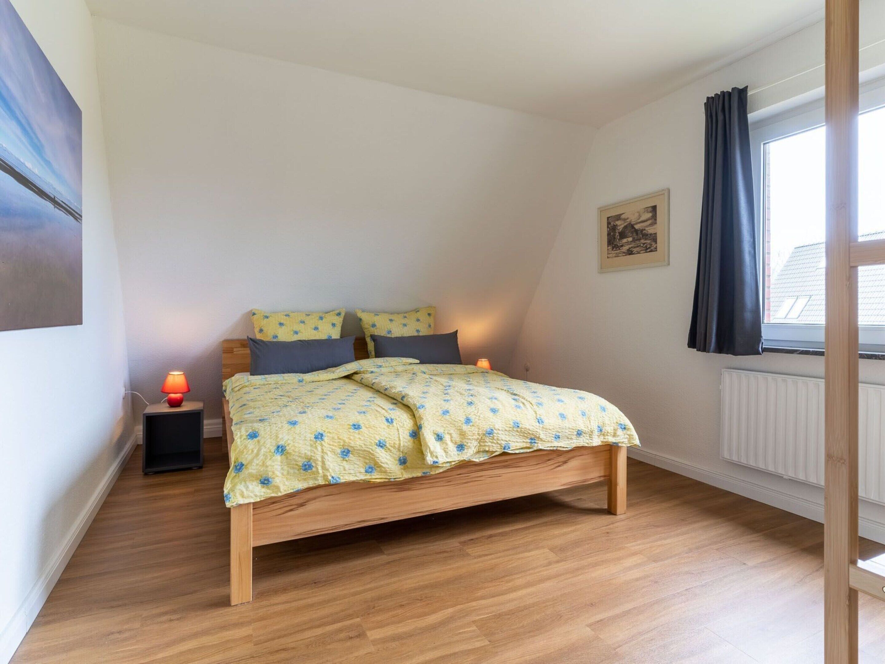 A bedroom with a wooden bed, a nightstand with a lamp, a window with curtains, and a radiator.