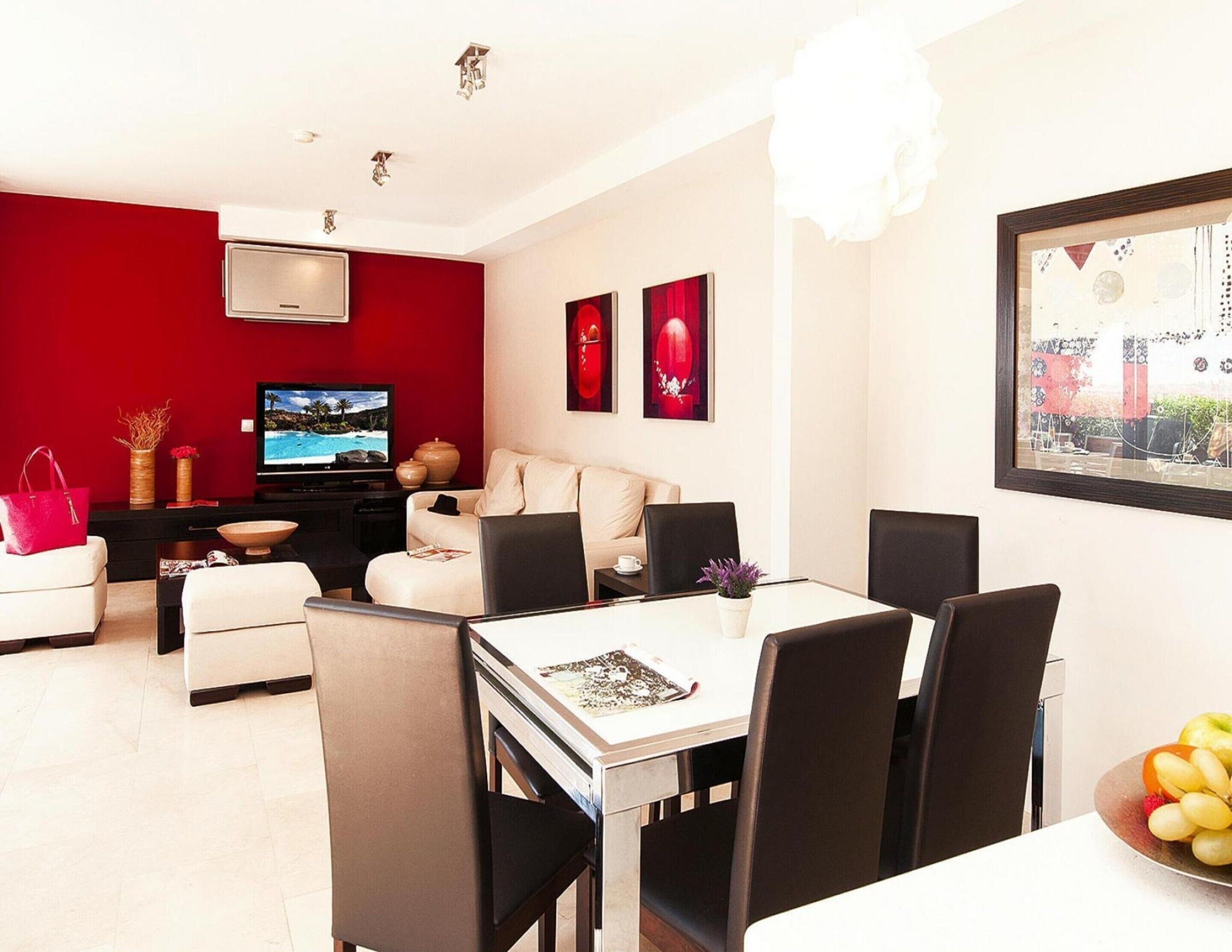A modern living room with a red accent wall, a flat-screen TV, a dining area with a white table and black chairs, and a large window with a view of a building.