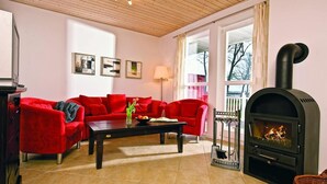 A living room with a red sofa set, a black wood-burning stove, and a window with a view of trees. - Appartements im Schlosspark Bad Saarow (Bad Saarow)