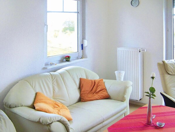 A white sofa with orange cushions, a small table with a vase and candle, and a window with a view of greenery. - Ferienhaus am Kummerower See, Kummerow (Kummerow)