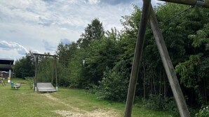 Puinen keinuteline vihreällä nurmialueella, taustalla on penkki ja katettu rakennelma.