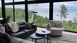 A modern living room with a large window, a telescope, and a view of a forest and lake. - Le Lord, River view, trail and snowmobile (Les Éboulements)