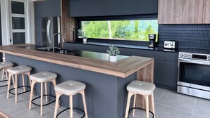 A modern kitchen with a central island, bar stools, a built-in oven, and a window with a view of greenery. - Le Lord, River view, trail and snowmobile (Les Éboulements)