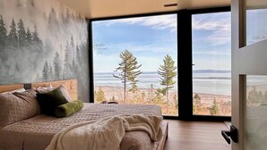 A bedroom with a large bed, a view of trees and a body of water through a large window. - Le Lord, River view, trail and snowmobile (Les Éboulements)