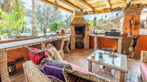 A rustic outdoor seating area with a brick fireplace, wicker furniture, and a view of the surrounding landscape. - Luxurious Private Cabin Pool Hottub & Fireplace (Alhaurin el Grande)