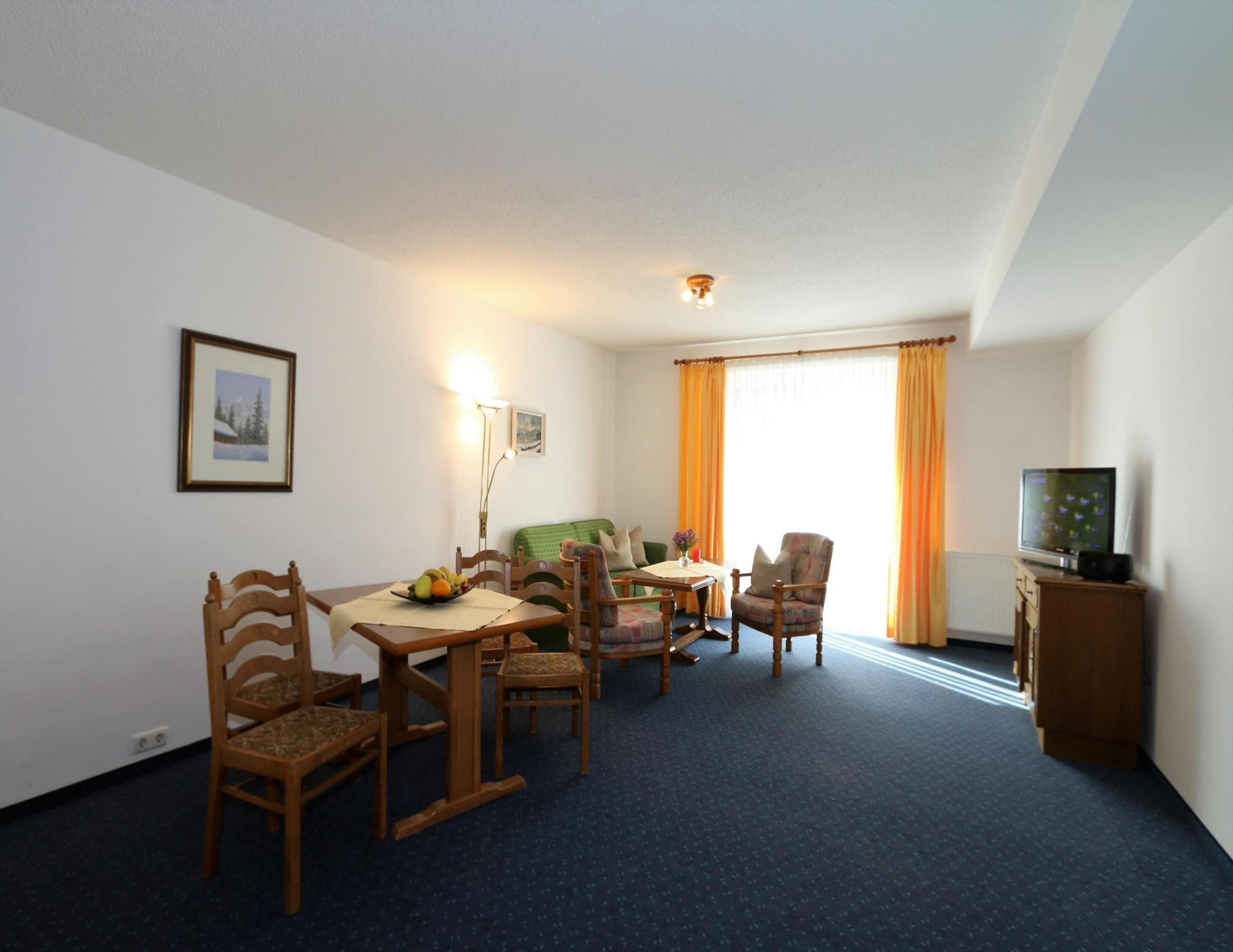 Habitación de hotel con una mesa de comedor, sillas, un sofá, un televisor y una ventana con cortinas.