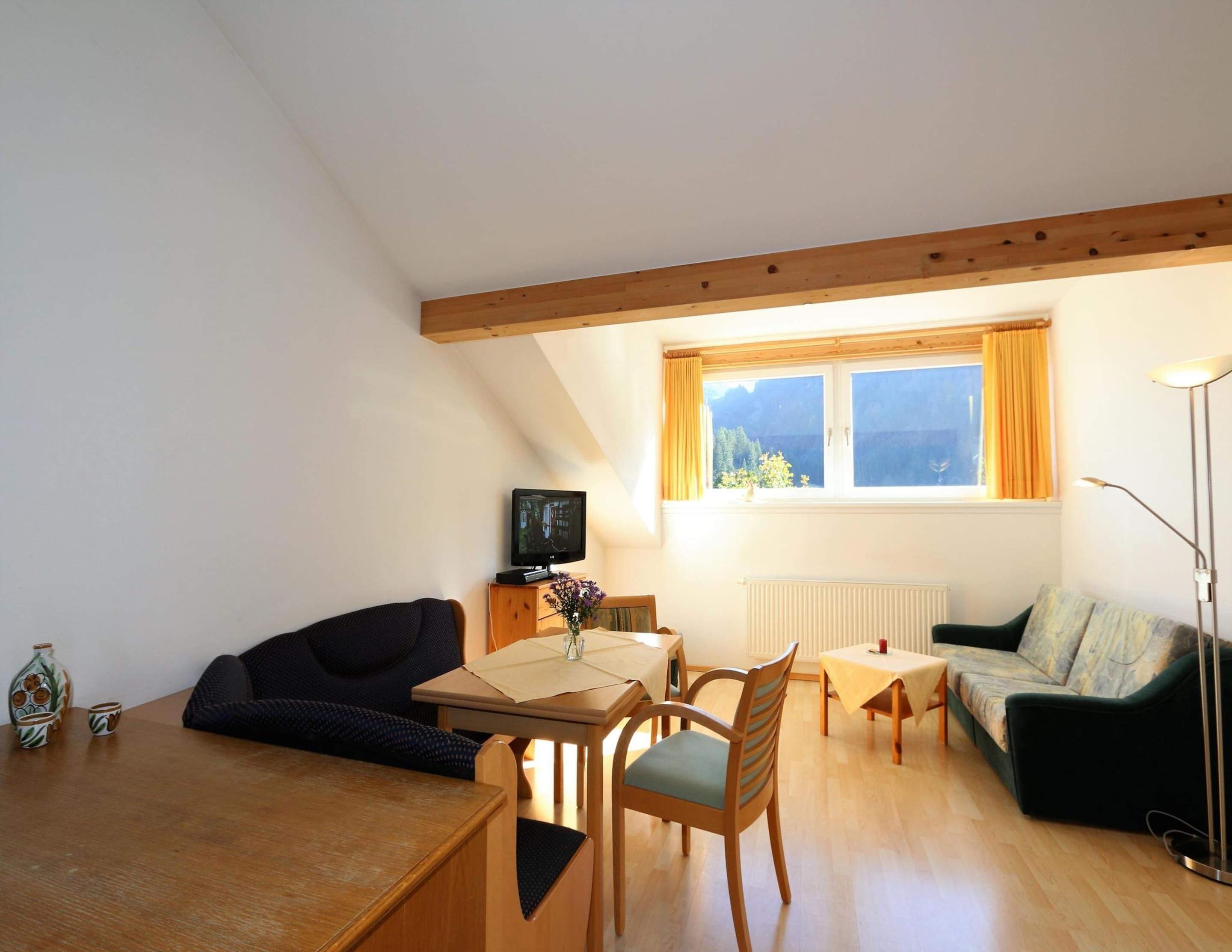 A cozy living room with a wooden ceiling, a TV, a sofa, a dining table, and a window with curtains.