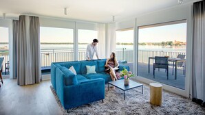 En rummelig opholdsstue med en stor blå sofa, et sofabord og udsigt over vand gennem store vinduer.