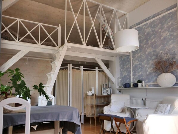 Una habitación acogedora con una cama tipo altillo, una mesa de comedor y una silla blanca. Se aprecia un ventanal con cortinas, una estantería blanca con adornos y una lámpara blanca.