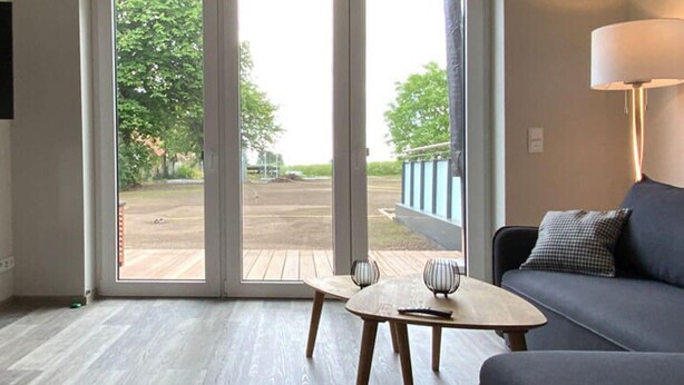 Ein modernes Wohnzimmer mit einer Couch, einem kleinen Tisch und einer Lampe. Eine Schiebeglastür führt in einen Außenbereich.