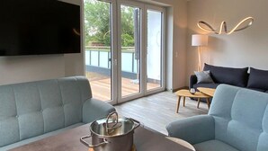 Ein modernes Wohnzimmer mit einer Couch, einem Couchtisch mit einer Pflanze, einem an der Wand montierten Fernseher und einer Glasschiebetür zu einem Außenbereich.
