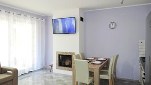 Un salon avec une cheminée, une table à manger avec des chaises, un canapé, un téléviseur fixé au mur et une horloge au mur.