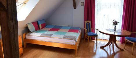 A bedroom with a sloped ceiling, a bed with a colorful quilt, a wooden chair, a small round table with a vase, and a window with red curtains.