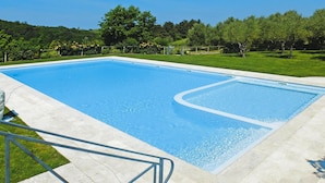 Une piscine rectangulaire à l’eau d’un bleu limpide, entourée d’une terrasse dallée et d’une zone herbeuse agrémentée d’arbres.