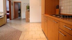 A modern kitchen with wooden cabinets, a stove, and a tiled backsplash. There is a hallway leading to a bedroom with a bed and a door. - Appartementhaus Nicole, Zinnowitz (Zinnowitz)