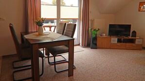 A dining area with a table, chairs, and a vase of flowers. There is a television on a wooden cabinet and a window with curtains. - Appartementhaus Nicole, Zinnowitz (Zinnowitz)