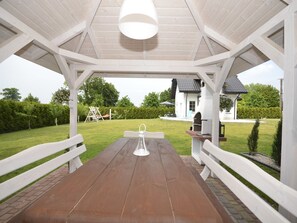 Une table de pique-nique en bois, placée sous une pergola blanche, offre une vue sur une maison et sur une balançoire en arrière-plan.