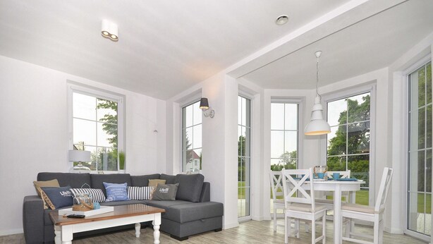 A spacious living room with a grey sofa, a wooden coffee table, and a white dining table with chairs. Large windows provide natural light and views of the outdoors.