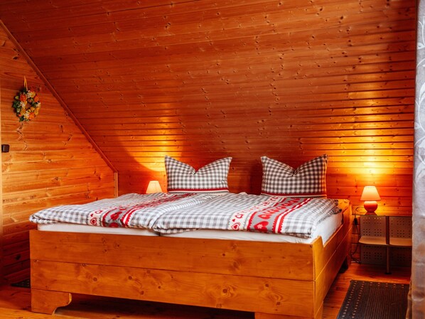 A wooden cabin bedroom with a sloped ceiling, a bed with checkered bedding, two bedside lamps, and a small table with a chair.