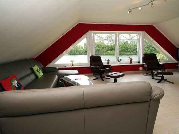 A modern living room with a sloped ceiling, a sofa, two armchairs, a small table, and large windows overlooking a lake. - Appartementhaus am Kiessee, Westerholt (Nenndorf)