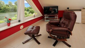 A living room with a brown leather recliner, a small table, a TV on a stand, and a window with a view of greenery. - Appartementhaus am Kiessee, Westerholt (Nenndorf)