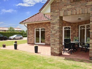A brick house with a tiled roof, a covered patio with a table and chairs, and a lawn with potted plants. - Appartementhaus am Kiessee, Westerholt (Nenndorf)