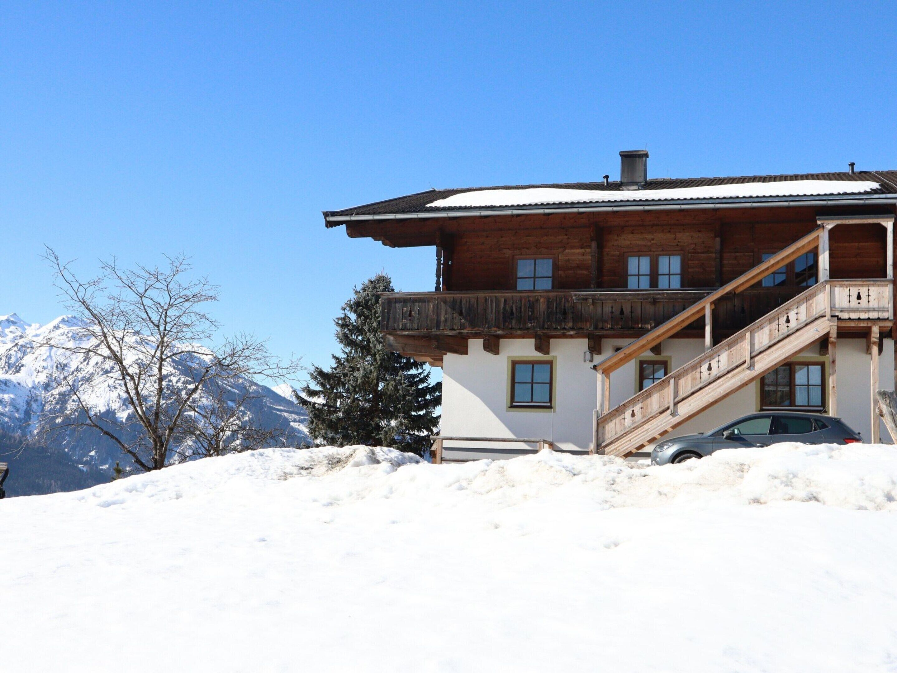 A wooden chalet with a balcony, a staircase, and a car partially buried in snow, set against a snowy mountain backdrop.