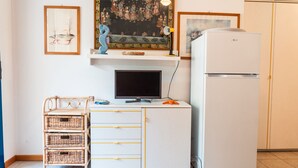 A compact kitchenette with a white refrigerator, a flat-screen TV, and a wicker storage unit. - Condominio Vera Cruz in Bibione-ex TUI (San Michele al Tagliamento)
