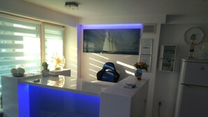 Une cuisine moderne avec un plan de travail blanc, un bar illuminé de lumière bleue, un tableau représentant un voilier et un réfrigérateur.