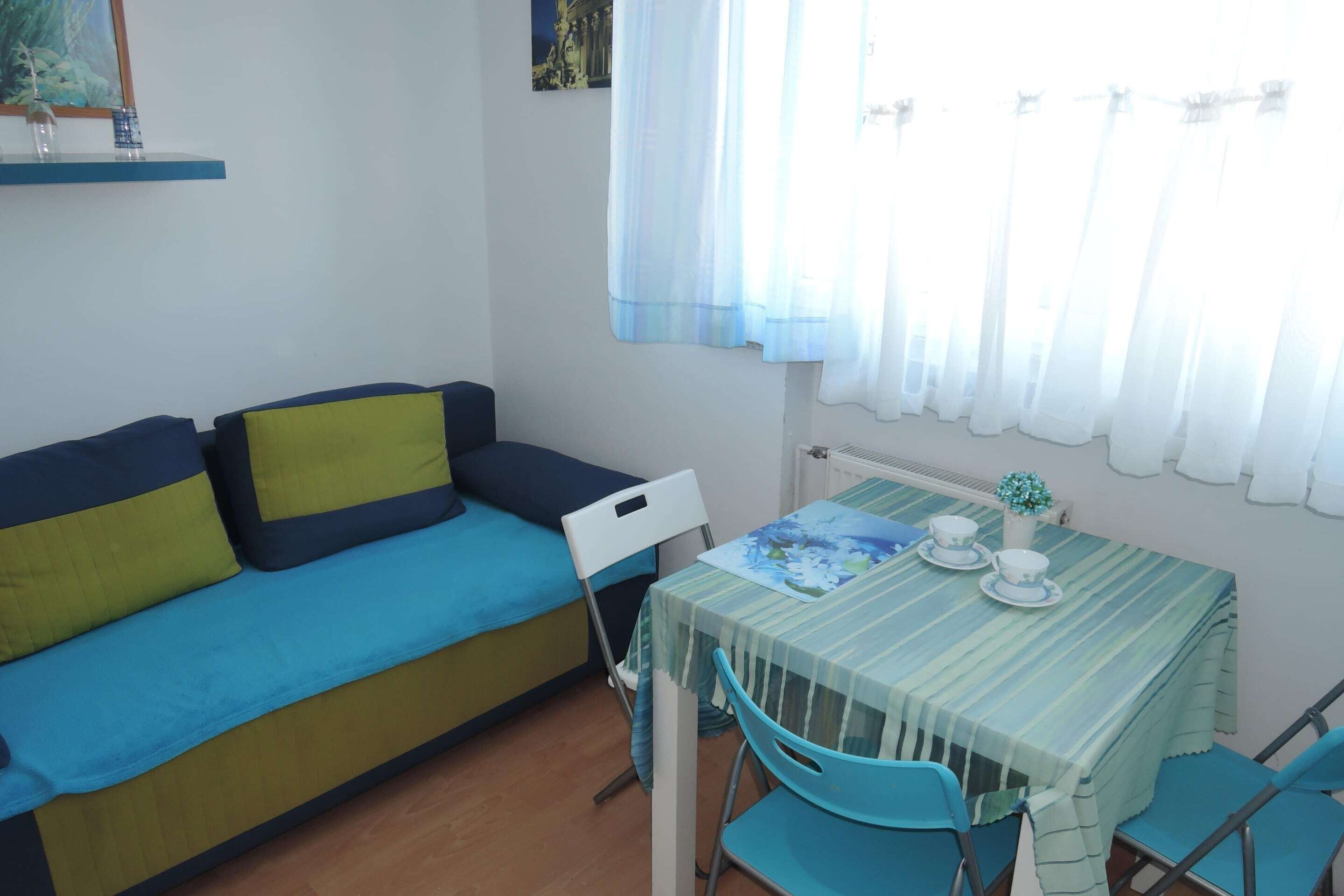 Une pièce avec un canapé bleu et vert, une chaise blanche, une table recouverte d’une nappe à rayures et une fenêtre ornée de rideaux légers.