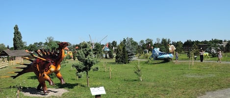 A dinosaur statue in a park with a sign, trees, and a clear sky.