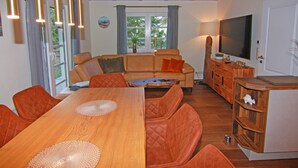 A cozy living room with a wooden dining table, orange chairs, a sofa, and a TV. - Doppelhaushalfte Dunenhaus II, Trassenheide (Trassenheide)