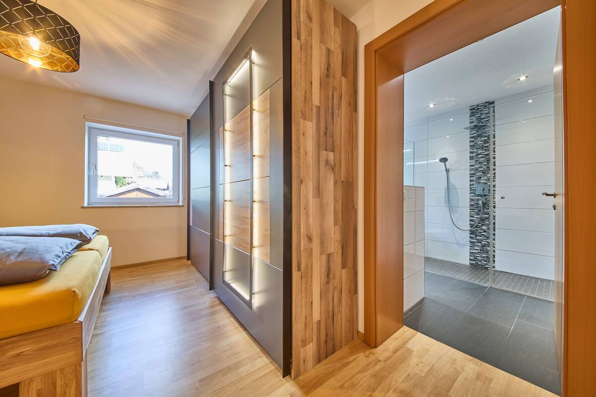 A modern bathroom with a shower, a wooden bench, and a window providing natural light.