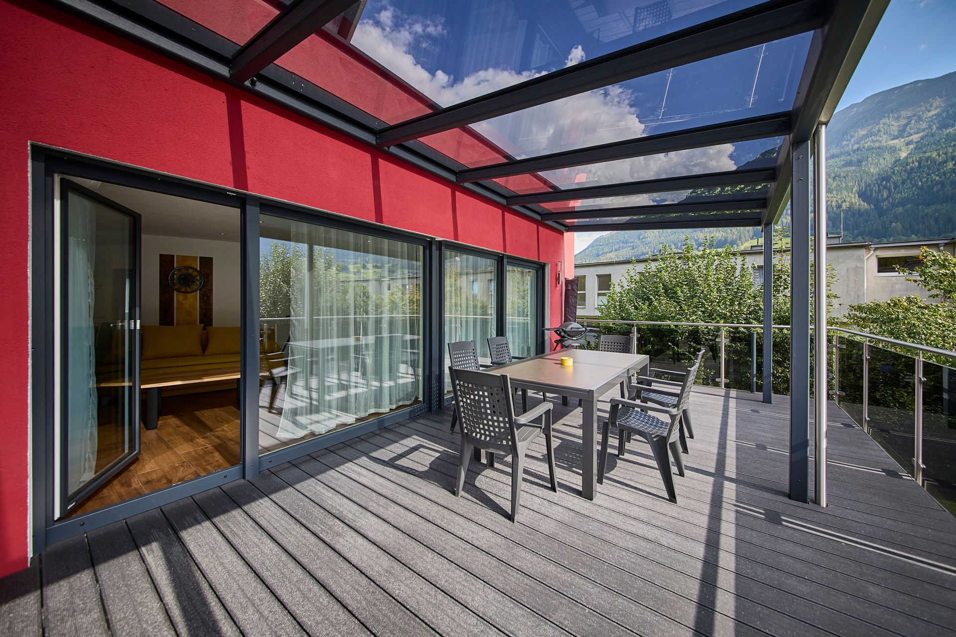 A modern outdoor deck with a glass door leading to a room, a table with chairs, and a view of a mountainous landscape.
