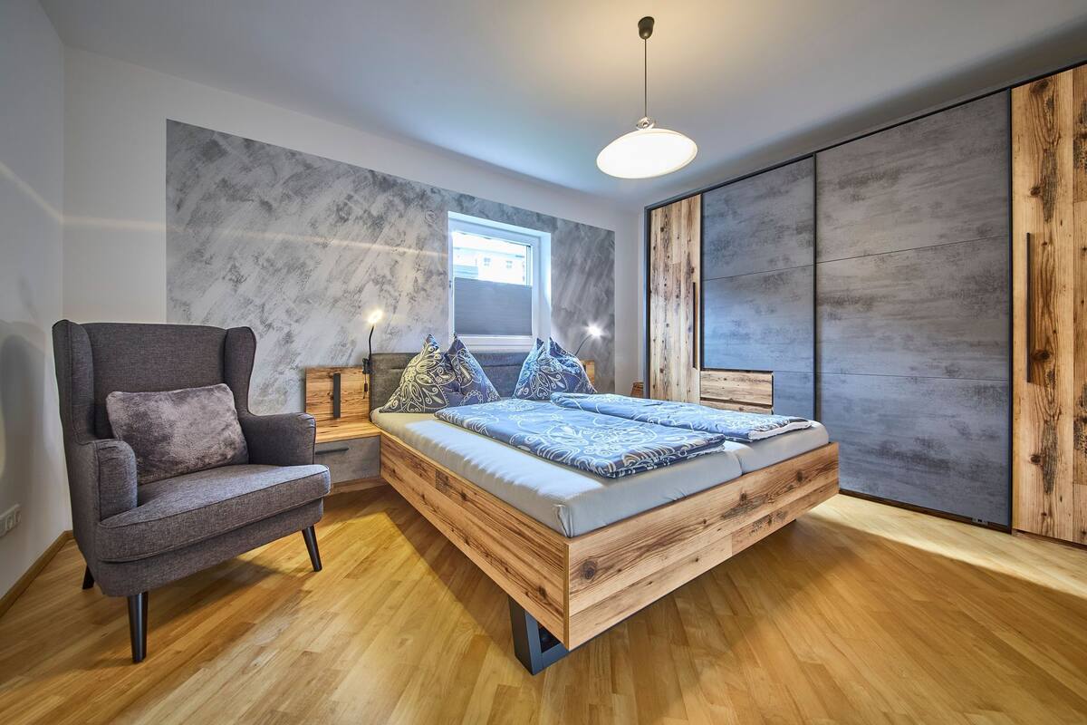 A modern bedroom with a wooden bed, a grey armchair, and a large window.