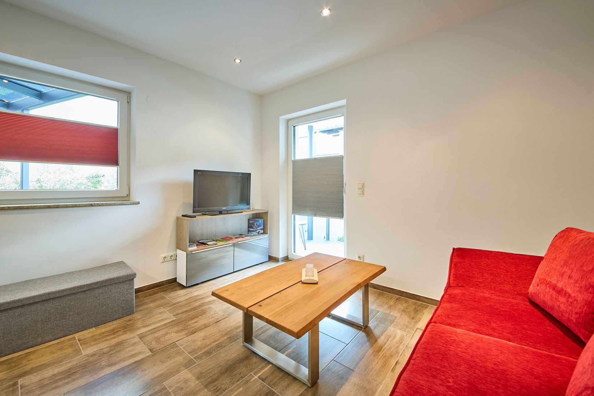 Un salón moderno con una mesa de madera, un sofá rojo, un televisor sobre un mueble de madera y una ventana con persianas.