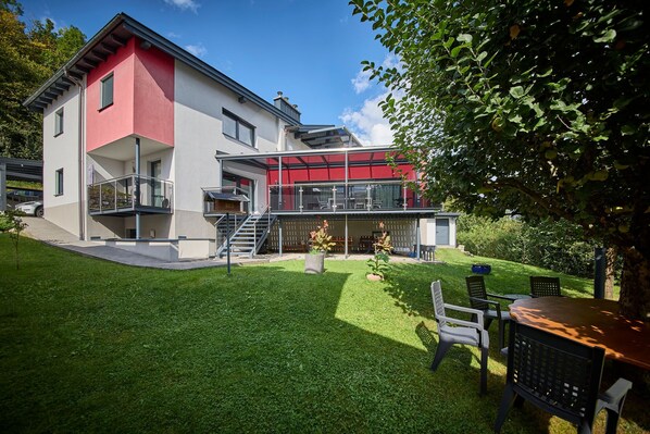 Una casa moderna de dos pisos con fachada roja y blanca, un invernadero de vidrio y un césped cuidado con mobiliario exterior.
