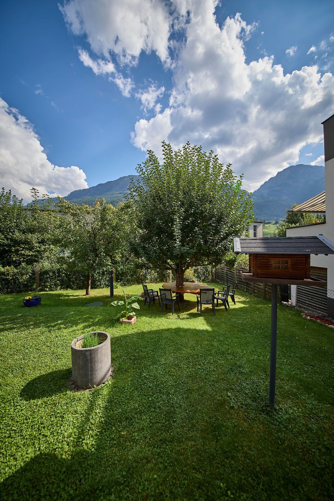 A well-maintained garden with a large tree, a wooden table with chairs, and a stone planter.
