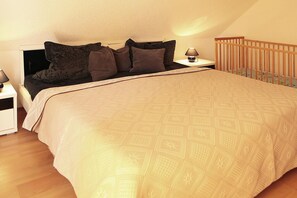 A bedroom with a large bed, a nightstand, a lamp, and a wooden crib. - Ferienhauser am Kummerower See, Verchen (Verchen)
